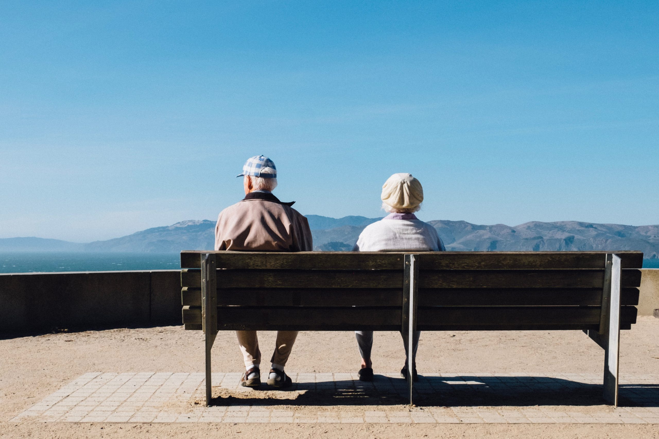 Un signore e una signora di spalle sono seduti su una panchina mentre guardano le colline accarezzate dal mare.